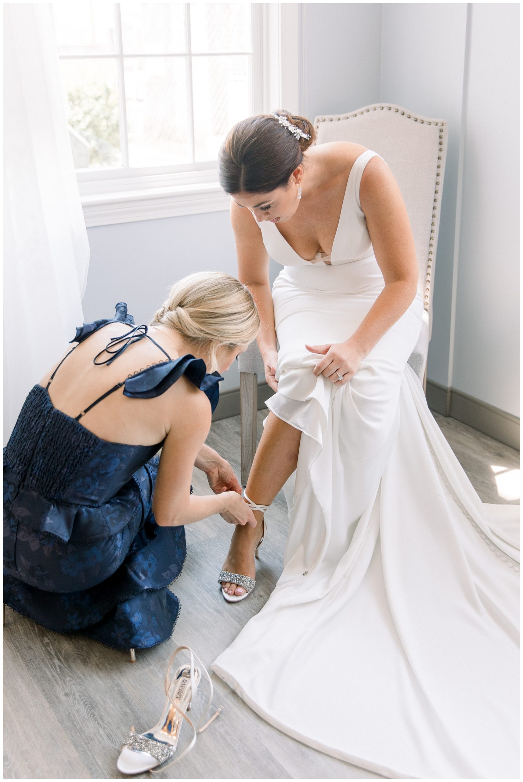 bridesmaid in navy gown helps bride with shoes