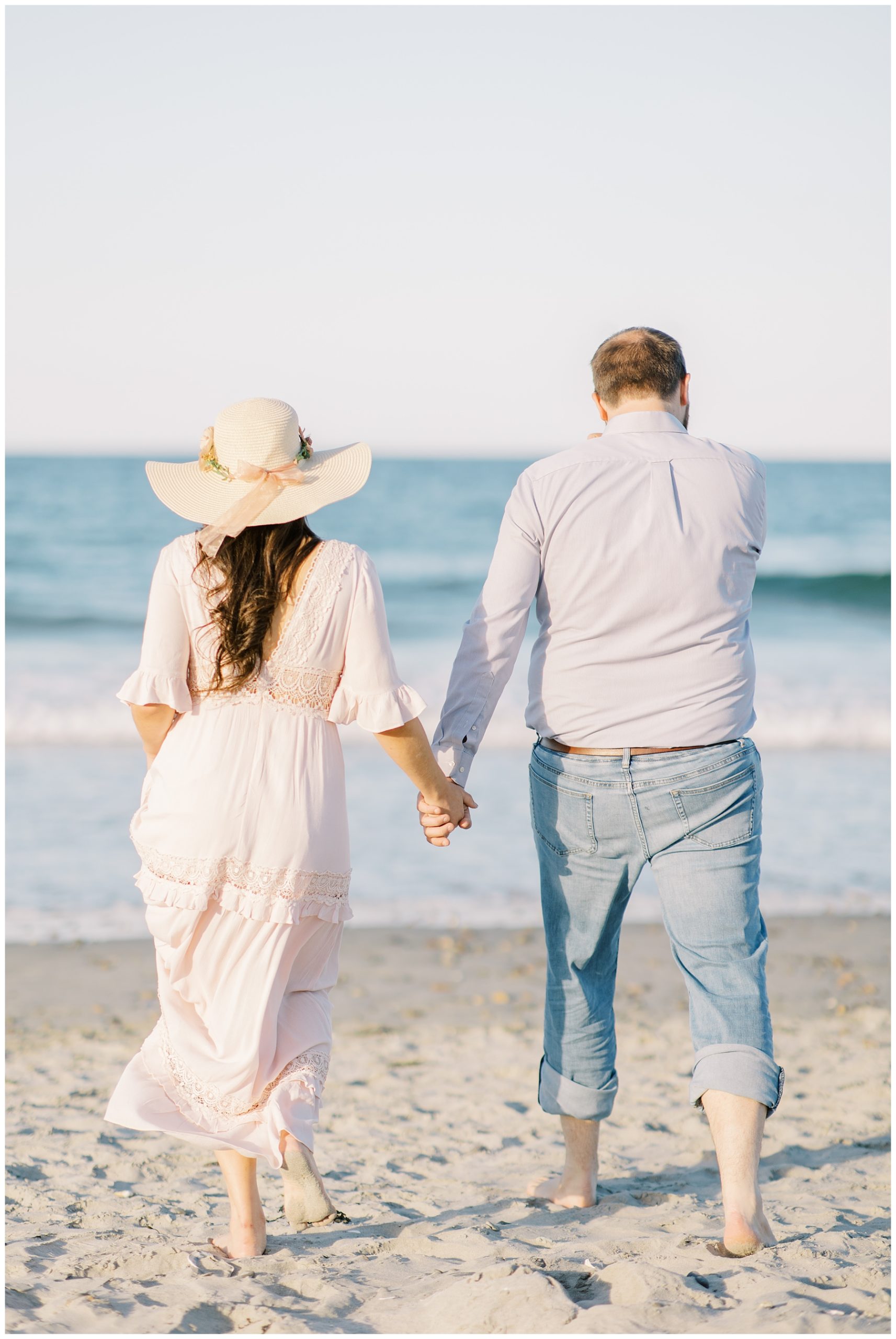woman in sun hat and dress hold her fiance's hand as they walk towards the ocean 