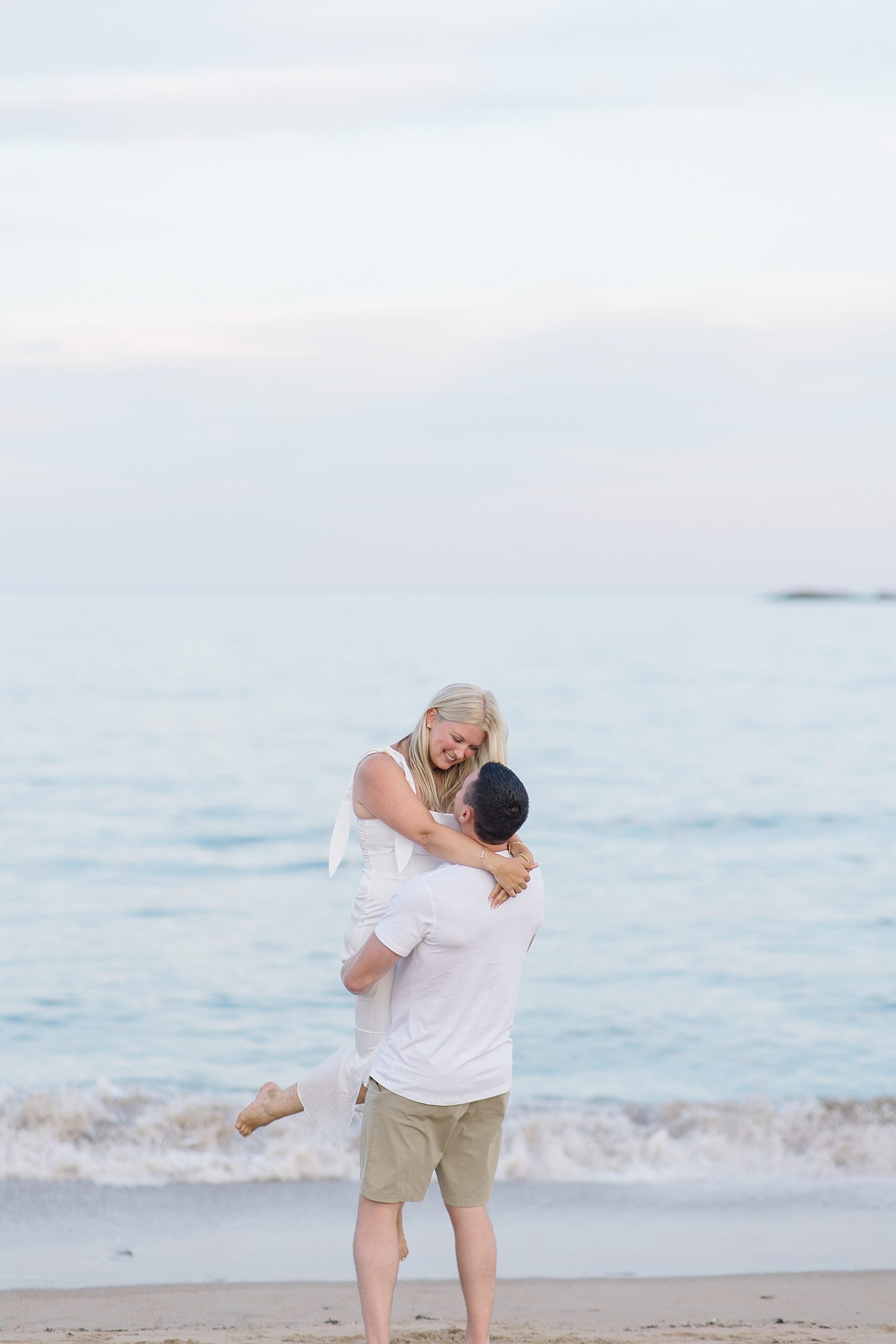 man lifts his fiancé up by the shoreline