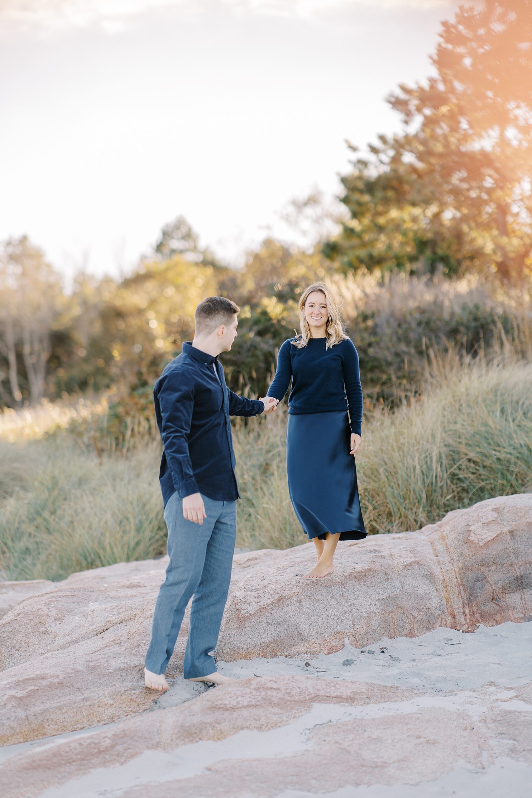 guy helps fiance walk over rocks on the beach