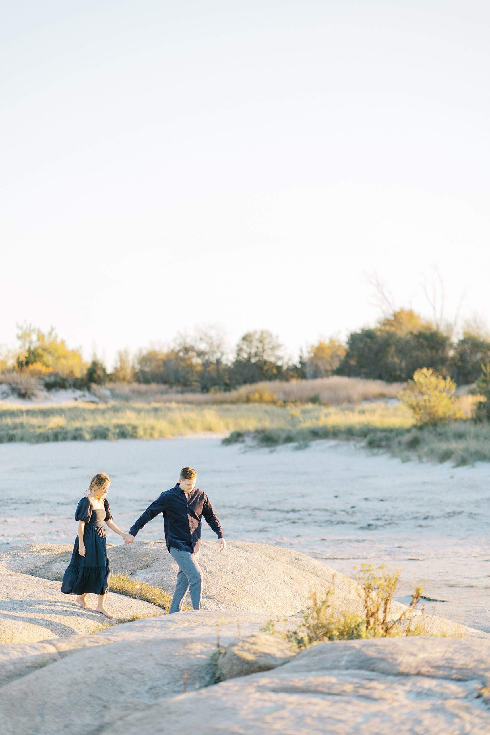 couple hold hands walking across rocky beach