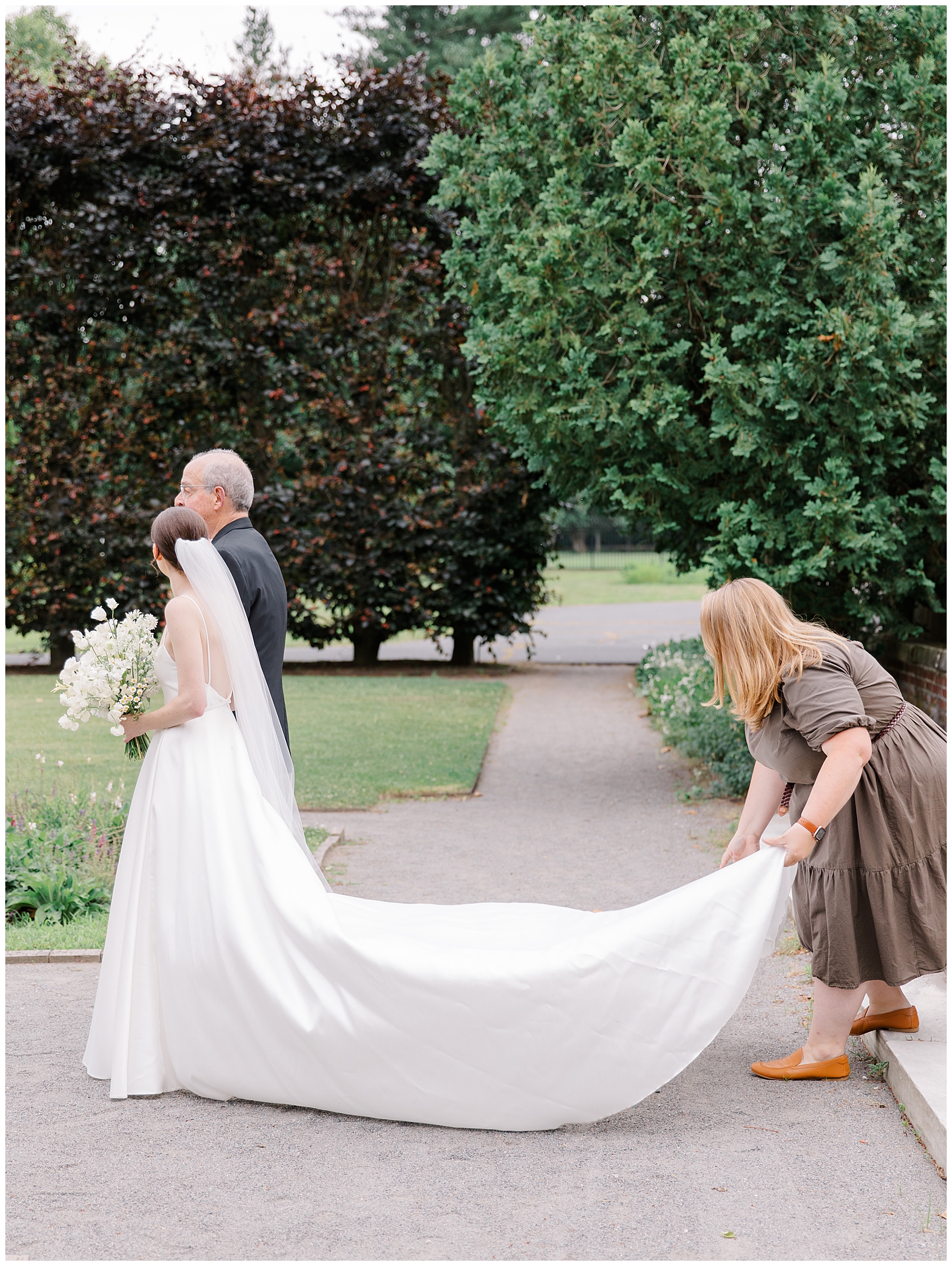 woman straightens bride's wedding dress as she walks down the aisle 