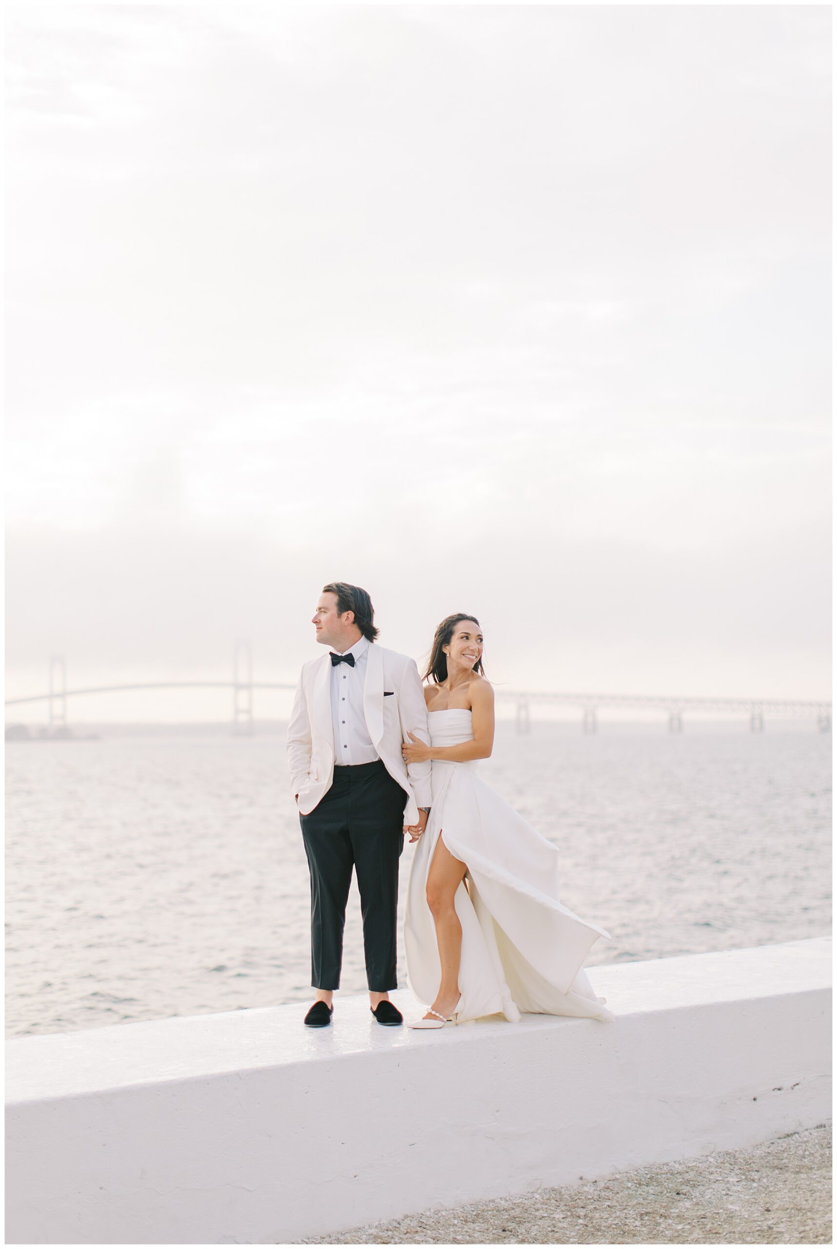 newlyweds stand on white wall by ocean in Newport from Coastal Wedding at Belle Mer
