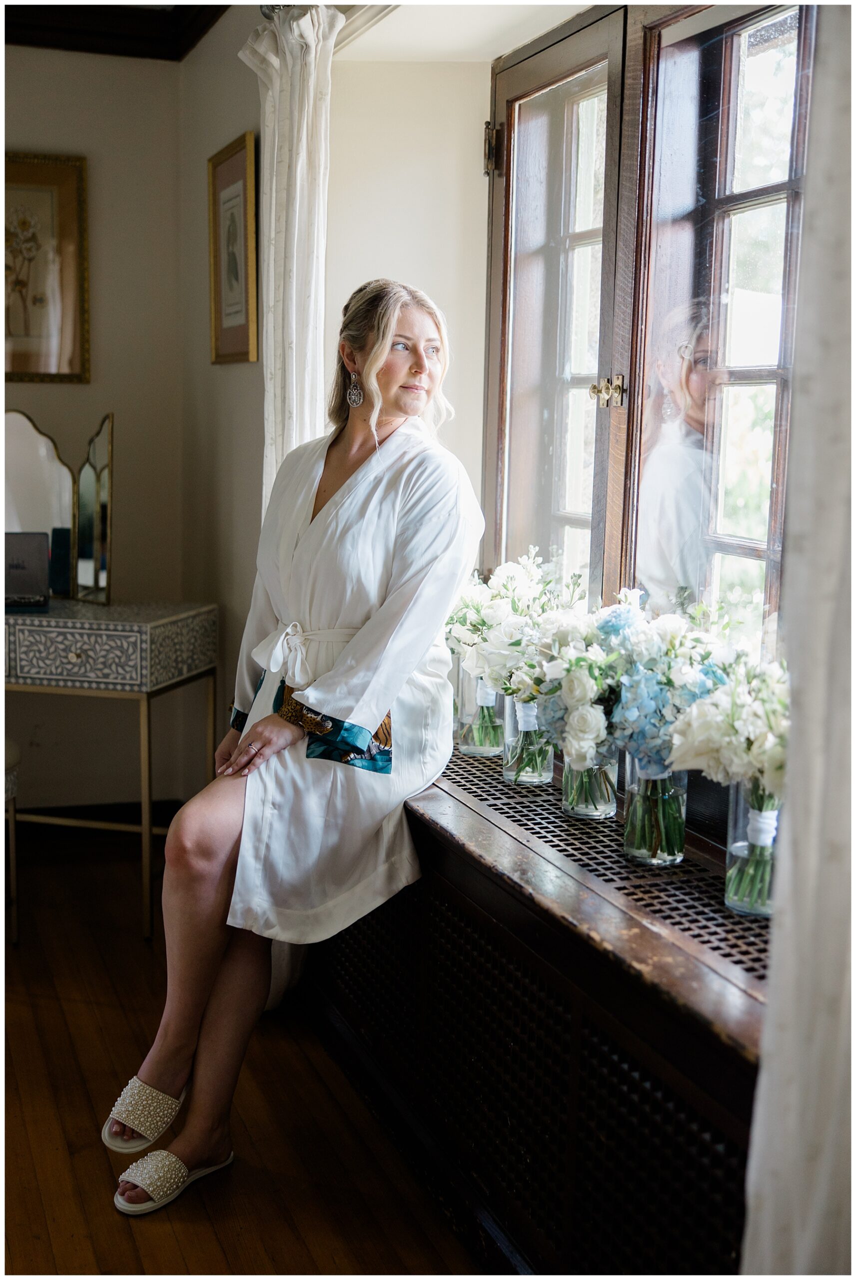 bride in white robe looking out window.