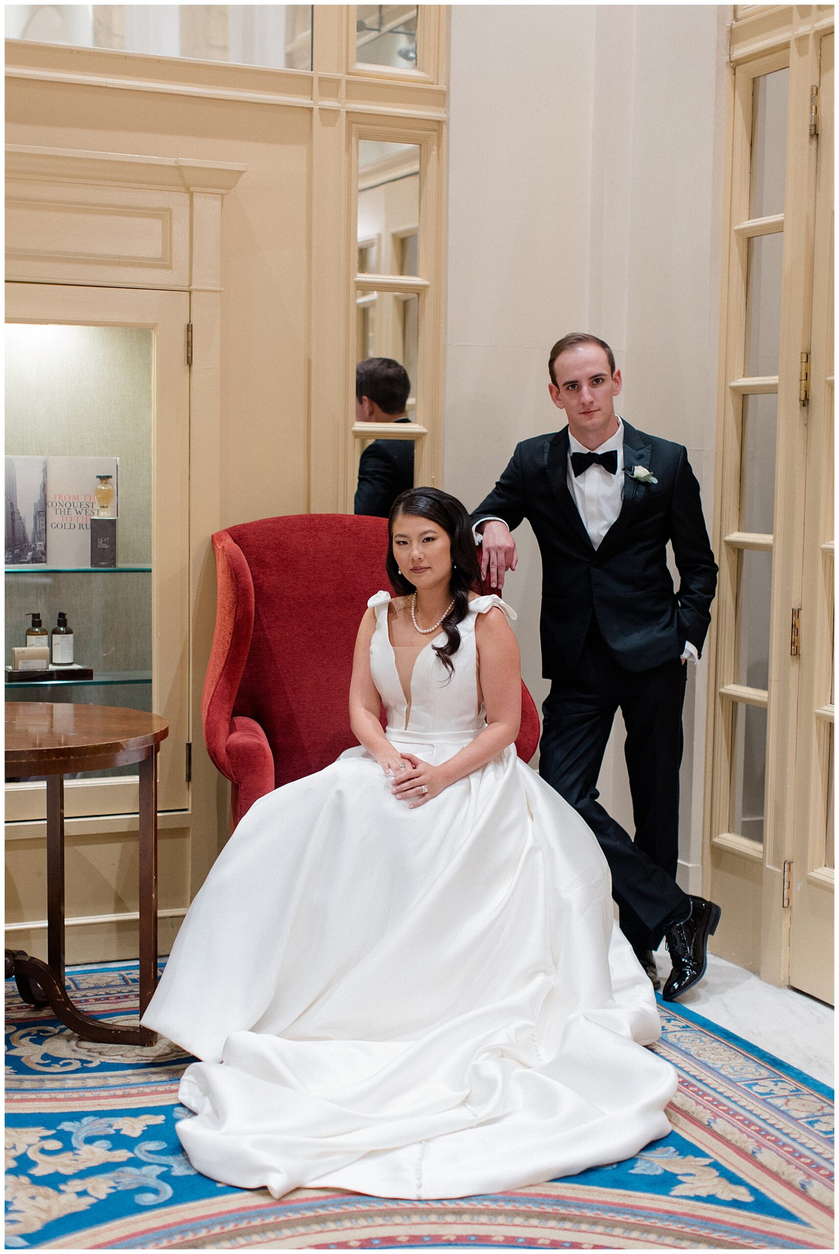bride sits in red chair with groom leaning against the back of the chair