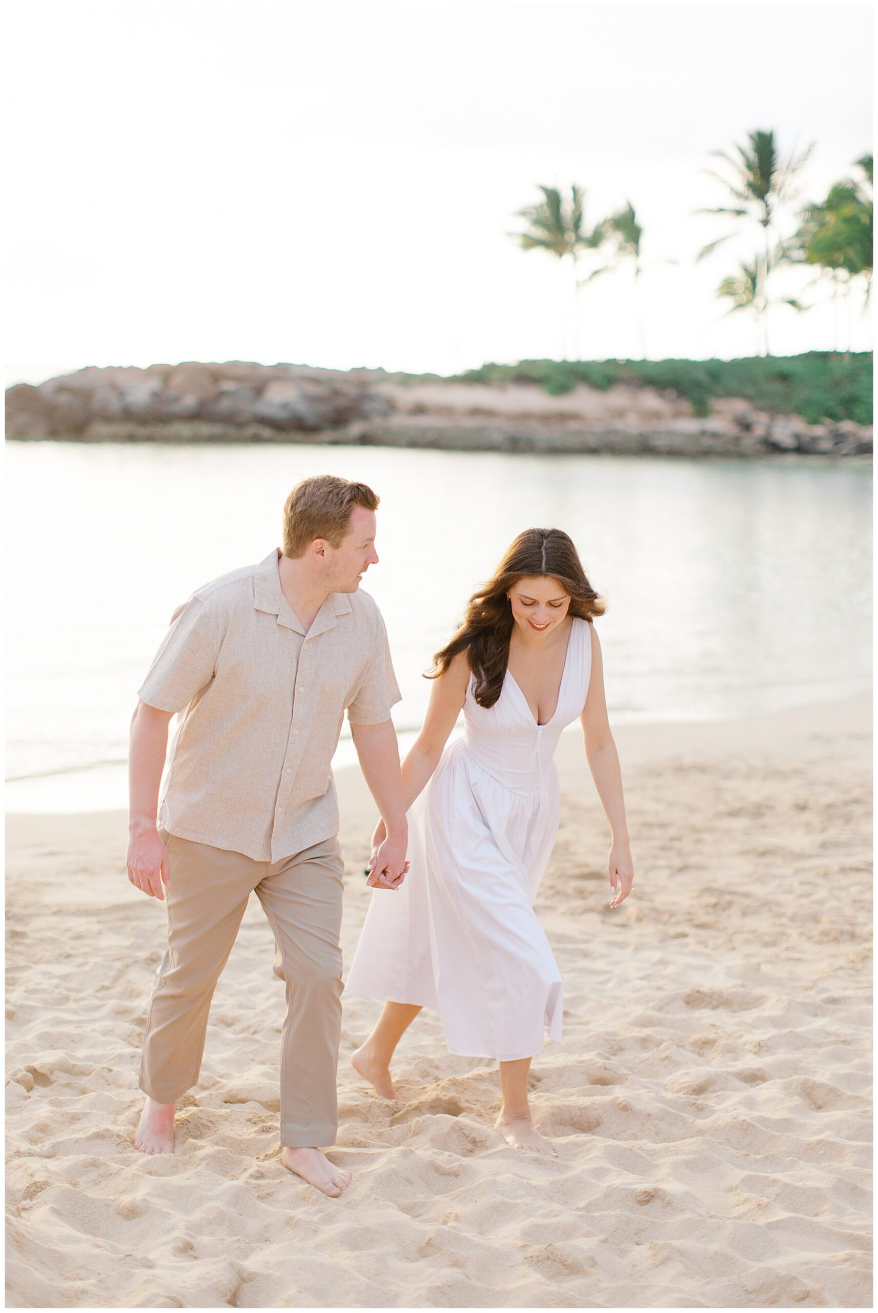 woman in white dress holds hands with fiance as they walk across the beach in Hawaii